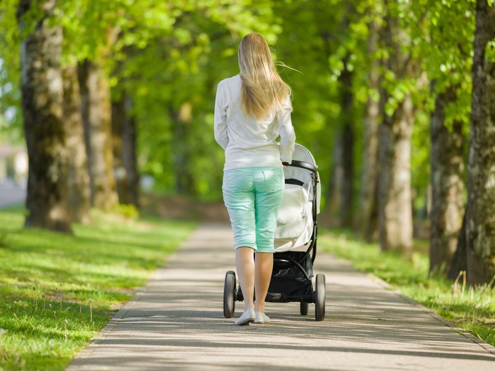 0 One young mother pushing white baby stroller and slowly walking through alley of trees in warm sunn