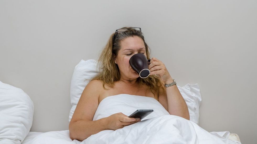 2022/07/1_Woman-in-her-hotel-bed-drinking-coffee-while-looking-at-her-mobile-phone-during-day.jpg