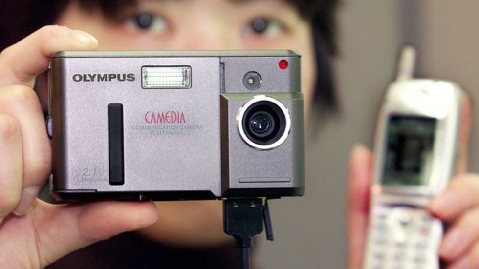 An out-of-focus person holds an Olympus Camedia camera in one hand, an early blocky mobile phone in another, with a cable running between them