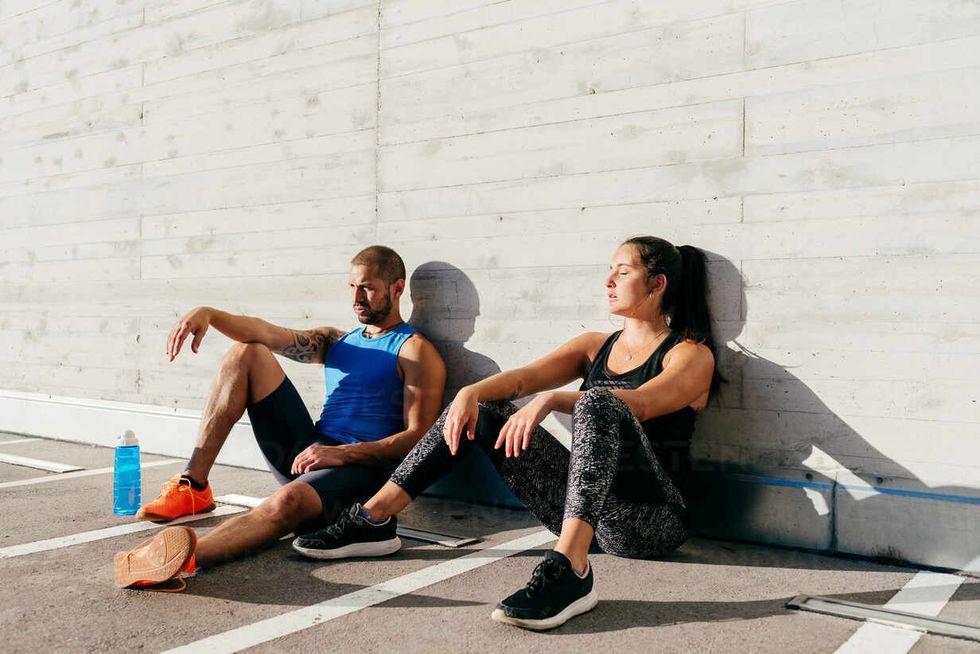 couple of exhausted athletes in sportswear sitting on street and leaning on stone wall while resting after intense workout on sunny day in city ADSF1