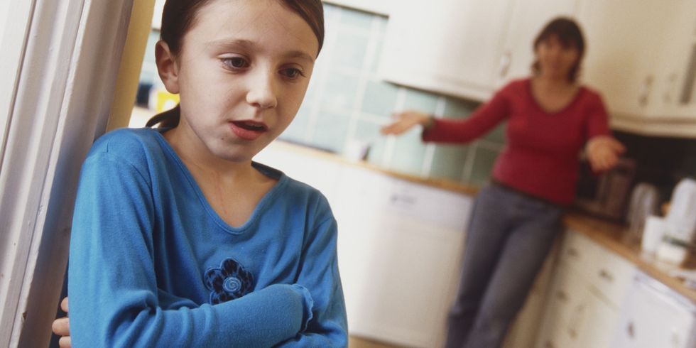 Girl standing in doorway of kitchen, arms folded, woman standing behind, arms extended.