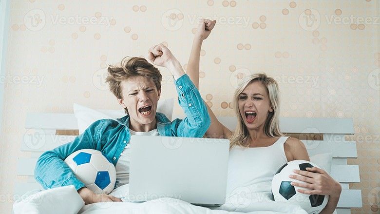 2022/11/happy-couple-watching-soccer-football-on-the-bed-photo.jpg