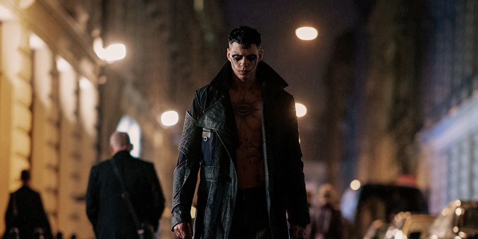 Një burrë me grim dramatik të fytyrës dhe një xhaketë lëkure të hapur ecën në një rrugë natën në The Crow (2024).