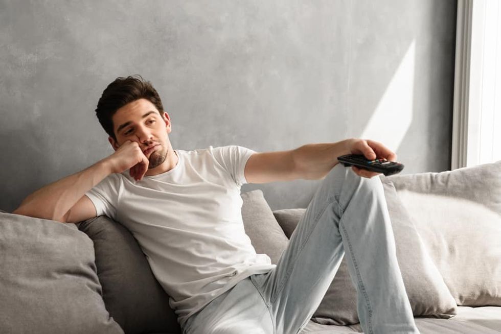 Photo of young bored man sitting on couch at home and changing TV channels with disinterest