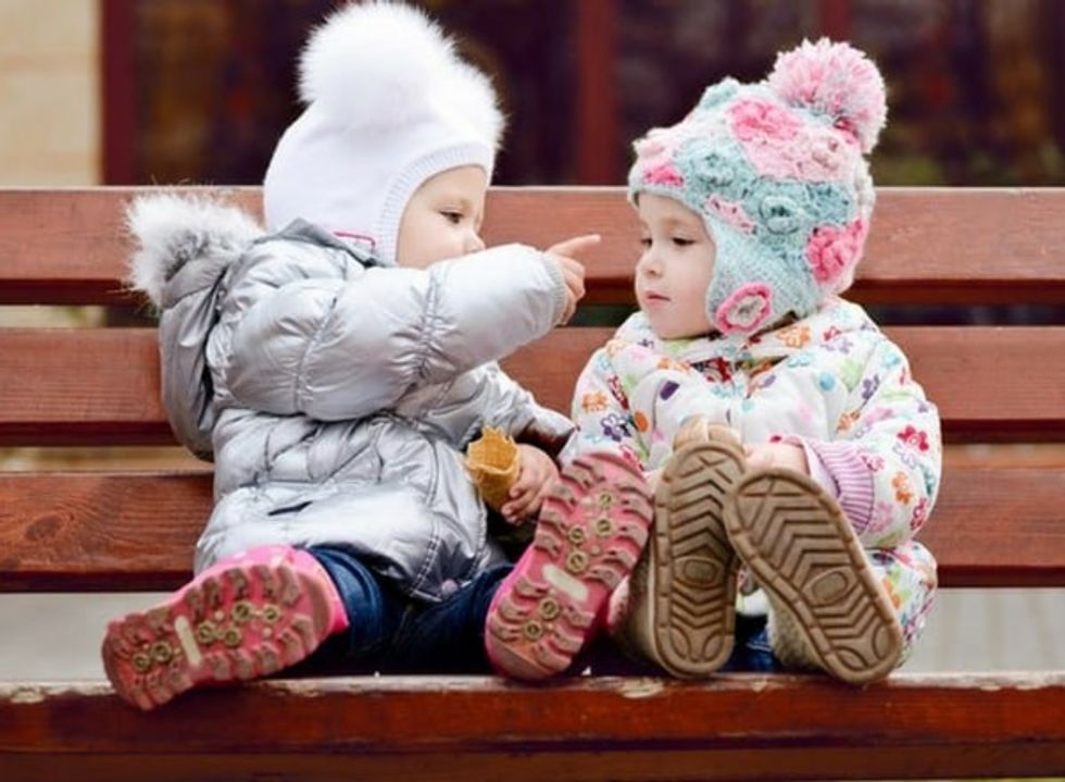 two babies in winter clothes talking and cooing with each other on a bench min