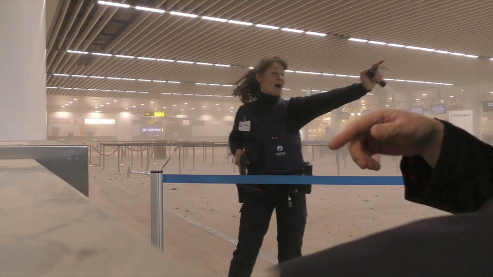 In this photo provided by Ralph Usbeck a police officers directs passengers in a smoke filled terminal at Brussels Airport, in Brussels after explosions Tuesday, March 22, 2016. Authorities locked down the Belgian capital on Tuesday after explosions rocked the Brussels airport and subway system, killing  a number of people and injuring many more. Belgium raised its terror alert to its highest level, diverting arriving planes and trains and ordering people to stay where they were. Airports across Europe tightened security.  (Ralph Usbeck via AP)