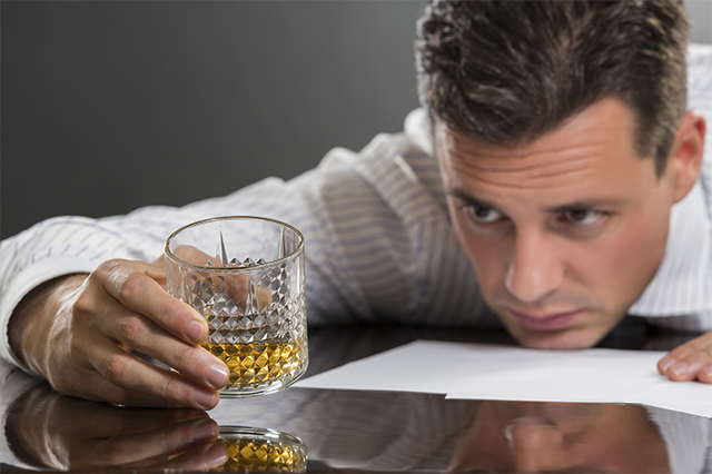 Portrait of sad frustrated young businessman resting his chin on a table while looking at the glass of alcohol in his hand. Selective focus on the glass.