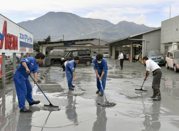 Auto dealership workers clean volcanic ash from the eruption of Mount Aso, back, in Aso city, Kumamoto prefecture, on the southern Japanese main island of Kyushu, Saturday, Oct. 8, 2016. Mount Aso has sent huge plumes of gray smoke as high as 11 kilometers (6.8 miles) into the air in one of the volcano's biggest explosions in years. The Japan Meteorological Agency says early Saturday's explosion also blew off bits of volcanic rock and ash, and raised the alert level for the area, extending the entry ban from just around the volcanic mouth to the mountain itself. (Hiroko Harima/Kyodo News via AP)