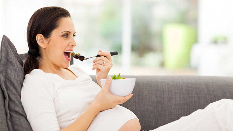 attractive pregnant woman eating vegetables on sofa